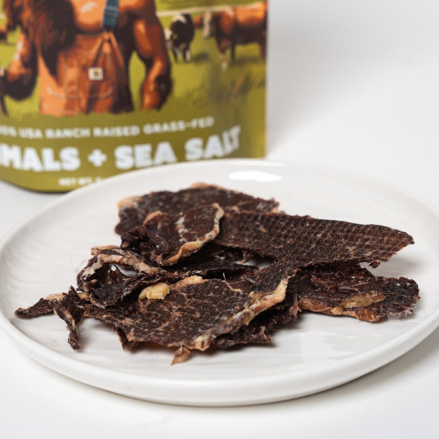 Dried beef jerky on a white plate with a packaging of 'MALS + SEA SALT' in the background.